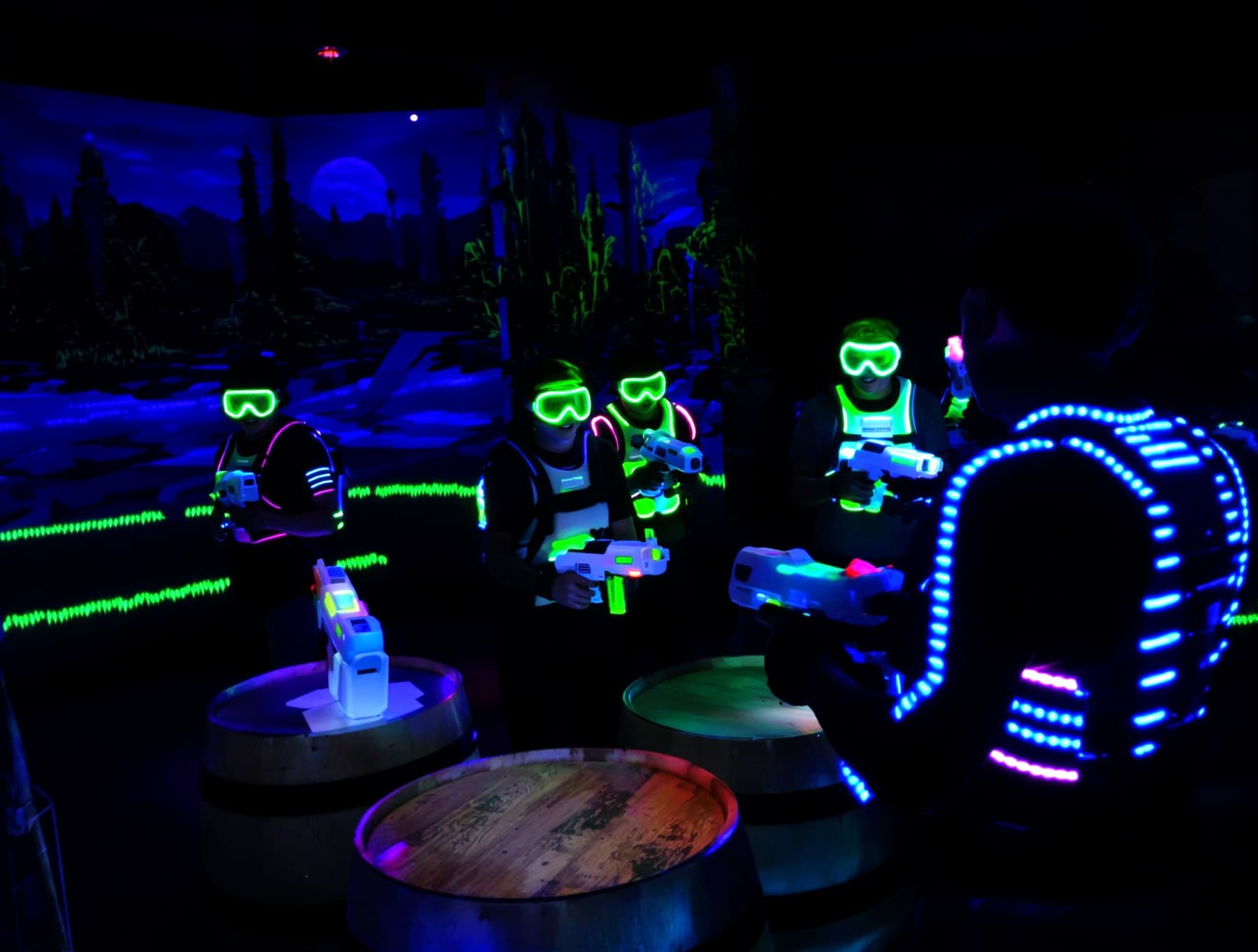 Players in goggle masks and glowing vests behind bunkers in a gel blaster arena